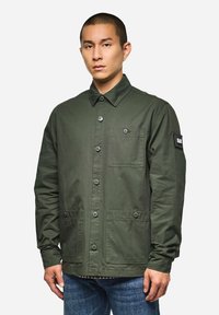 Grüne Baumwolljacke mit Knopfleiste, zwei Fronttaschen und einer Brusttasche. Hat einen umschlagbaren Kragen und ein Patch am Ärmel.