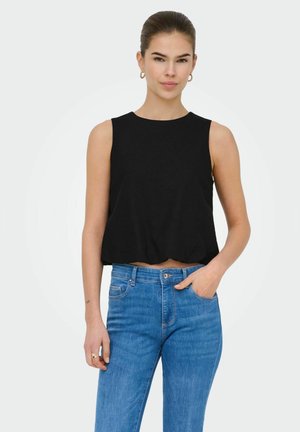 Jeune femme portant un crop top noir sans manches et un jean bleu taille haute, une main dans la poche, sur un fond blanc uni.