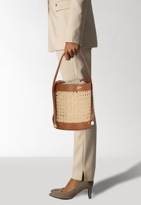 Sac seau avec un corps tissé beige, des accents en cuir marron, une bandoulière réglable et une fermeture à cordon en haut.