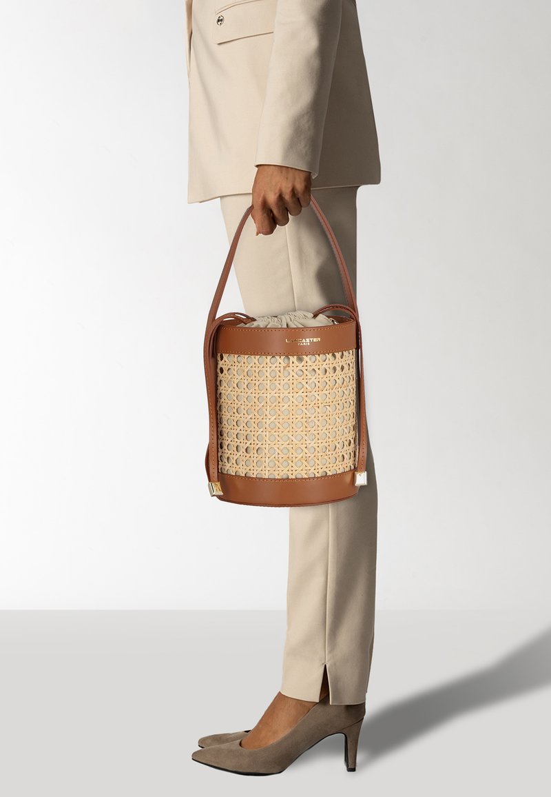 Sac seau avec un corps tissé beige, des accents en cuir marron, une bandoulière réglable et une fermeture à cordon en haut.