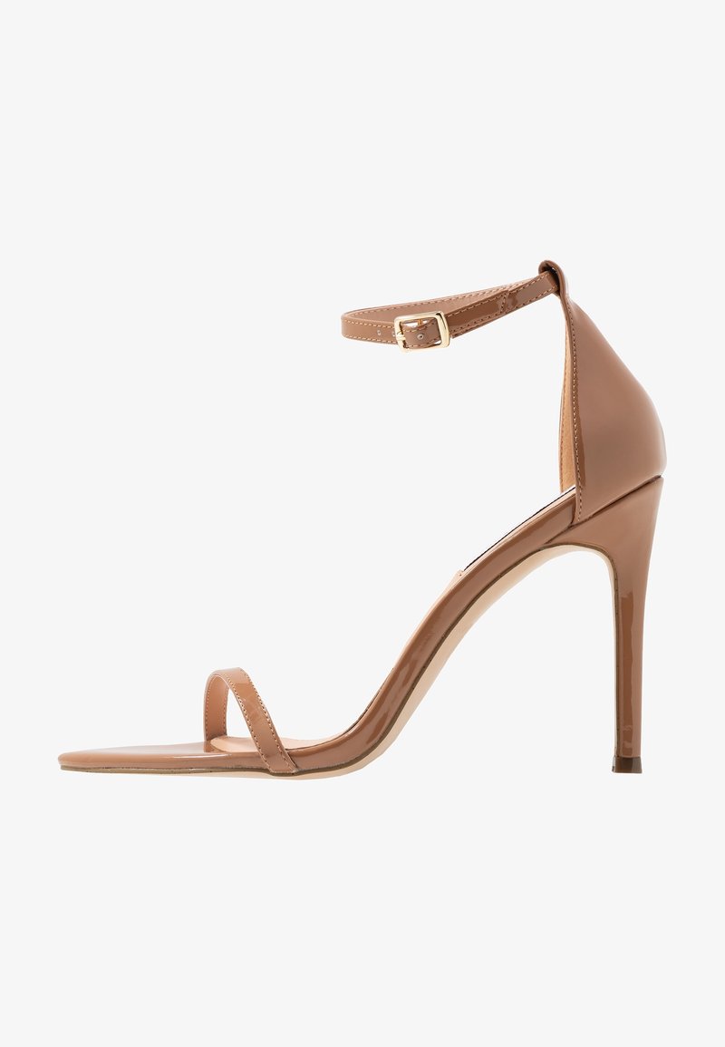 Sandale à talon haut de couleur beige avec une fine bride sur les orteils et une bride réglable autour de la cheville, dotée d'une petite boucle dorée, vue de profil.