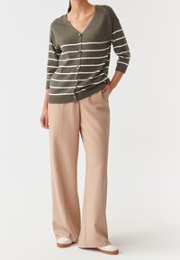 Kobieta ubrana w oliwkowo-zielony i biały prążkowany sweter na guziki, beżowe spodnie o szerokich nogawkach oraz białe tenisówki z brązowymi podeszwami.