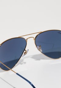 Zlaté kovové aviátor brýle se tmavě modrými skly. Průhledné nosníky a elegantní modrý stránek mají zakřivený design.