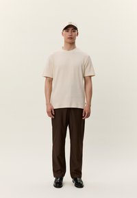 Jauns vīrietis stāv, valkājot bēša krāsas cepuri un t-kreklu, tumši brūnas bikses un melnus apavus uz vienveidīga gaiša fona.