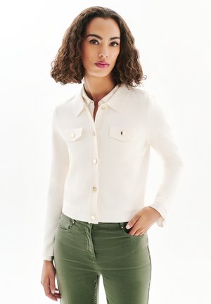 Kvinde med krøllet hår iført en hvid jakke med knapper og grønne bukser, stående med den ene hånd i lommen, vendt fremad.