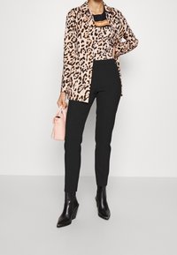 Długa koszula w cętki w odcieniach beżu i brązu, czarne dopasowane spodnie, czarne botki na obcasie oraz mała różowa torebka.