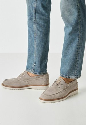 Grijze suède schoenen met een mocassinstijl ontwerp, voorzien van witte rubberen zolen, ronde veters en decoratieve stiksels, gecombineerd met een blauwe spijkerbroek.