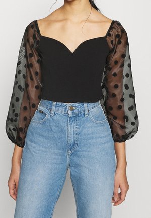 Femme portant un haut noir épaule dénudée avec des manches longues en tissu transparent à pois noirs et un jean bleu clair taille haute.