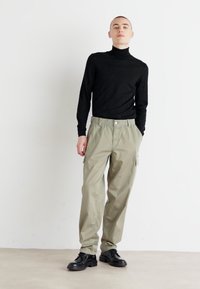 Jersey de cuello alto negro, pantalones cargo en un tono oliva claro y zapatos negros. Los pantalones tienen cintura elástica y bolsillos laterales, con una textura de tela suave.