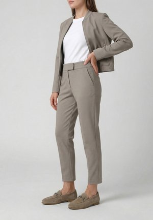 Frau trägt taupefarbenen taillierten Blazer und passende schmal zulaufende Hose mit beigen Loafers und weißem Rundhals-T-Shirt, Hand auf der Hüfte.