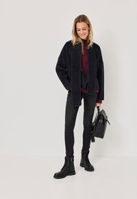 Čierna fleecová bunda so širokým golierom, vínový sveter, čierne úzke džínsy a čierne šnurovacie členkové topánky. Obsahuje čiernu kabelku.