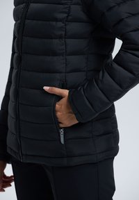 Hand in der mit einem Reißverschluss versehenen Tasche einer schwarzen, gesteppten Pufferjacke, die mit schwarzen Hosen auf einem hellgrauen Hintergrund getragen wird.