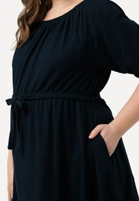 Tummansininen mekko, joka on valmistettu pehmeästä kankaasta ja jossa on olkaimeton pääntie, joustava vyötärö ja sidottava vyö. Sisältää sivutaskut.