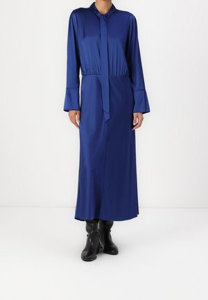Frau trägt ein langärmliges, königsblaues Midi-Kleid mit gebundenem Halsdetail und schwarzen Stiefeletten auf weißem Hintergrund.