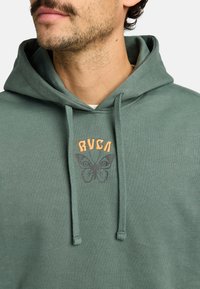 Felpa verde realizzata in tessuto morbido, con una grafica di una farfalla e il logo RVCA in arancione. Include lacci e una spaziosa tasca a marsupio.