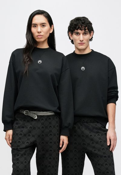 Fekete pulóverek kerek nyakkivágással, amelyeket két személy visel, központi logóval díszítve. Mindketten ugyanolyan mintás fekete nadrágot viselnek.