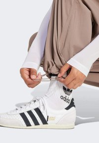 White athletic shoe with black stripes, beige tracksuit pants, and long white sleeves. Hands adjust elastic at pants' ankle, showing textured fabric.