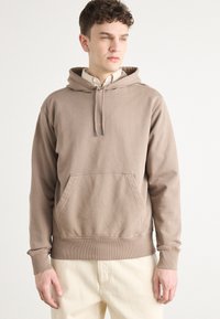 Bruine hoodie met een kangoeroezak, ribgebreide manchetten en zoom. Bevat een klein logo op de borst. Gedragen over een overhemd met kraag.