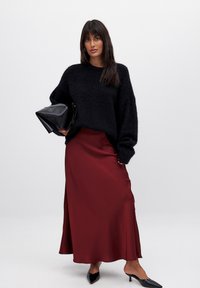 Schwarzer, flauschiger Strickpullover; burgunderfarbener Satinrock; schwarze spitze High Heels; schwarze dreieckige Handtasche; glatte, weiche Texturen.