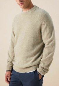Pull beige clair en tricot doux, avec un col rond côtelé, des manches longues et un ourlet côtelé. Porté sur un pantalon navy.