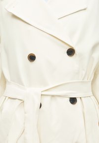 Manteau croisé couleur crème avec boutons ronds foncés et ceinture assortie en tissu nouée à la taille.