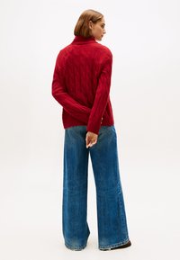 Suéter rojo de punto trenzado con cuello alto, combinado con unos vaqueros anchos de color azul con dobladillo deshilachado. El conjunto tiene un ajuste holgado.