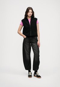 Svart fleece vest med glidelås foran, båret over en rosa t-skjorte. Mørke, vide denim bukser og svarte sneakers med hvite snører fullfører stilen.