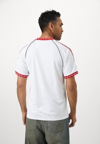 Vit sporttröja med röda detaljer på kragen och ärmarna, med ett strukturerat mönster på tyget. Avslappnad passform med rund halsringning.