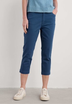 Person wearing navy blue cropped pants, light blue top, and white lace-up sneakers standing with one hand in pocket.