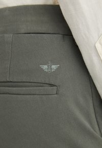 Olive green pants made of textured fabric, featuring a small logo with wings and a caduceus design above a back pocket detail.