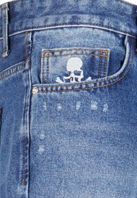 Jean en denim avec un délavage bleu clair, design classique à cinq poches et un crâne et des os blancs brodés sur la poche arrière.