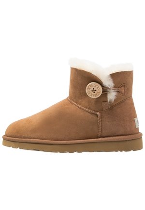 Brązowy zamszowy botek za kostkę z podszyciem z owczej wełny, okrągłym guzikiem z logo UGG i płaską gumową podeszwą.