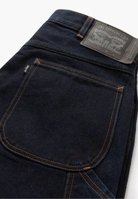 Mörka jeans i denim med orange sömmar, en bakficka och en grå läderetikett med varumärkesinformation vid midjan.