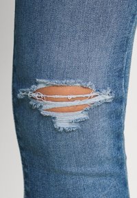 Pantalones vaqueros de denim con una zona de rodilla visiblemente desgastada y una gran rasgadura deshilachada. La tela es de un azul medio clásico con una textura sutil.