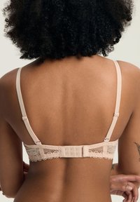 Kobieta z kręconymi włosami, ubrana w beżowy koronkowy biustonosz z tyłu, z regulowanymi ramiączkami i zapięciem na haftkę.