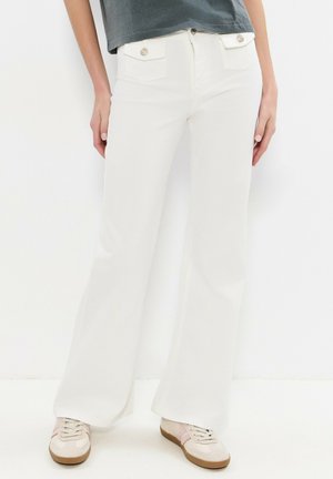 Personne portant un pantalon blanc à jambes larges avec des poches à rabat avant et des baskets beige, debout devant un fond blanc.