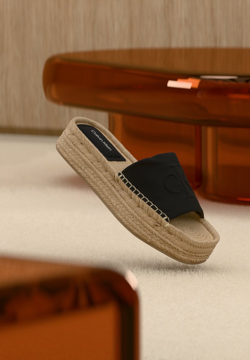 Sandale mule espadrille à plateforme beige avec une bride en tissu noir embossée du logo « CK », flottant à l'intérieur près de meubles de couleur ambre.