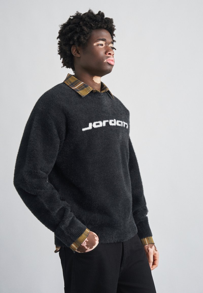 Μαύρο πουλόβερ με χνούδι και λευκό λογότυπο "Jordan", φορεμένο πάνω από καρό πουκάμισο με γιακά σε καφέ και κίτρινους τόνους. Τα μανίκια έχουν σχέδια ως διακοσμητικά στοιχεία.