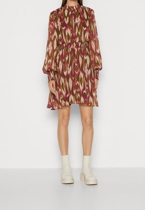 Kvinde iført en langærmet, knælængde kjole med brune, pink og beige abstrakte mønstre og hvide ankelstøvler, stående mod en hvid baggrund.