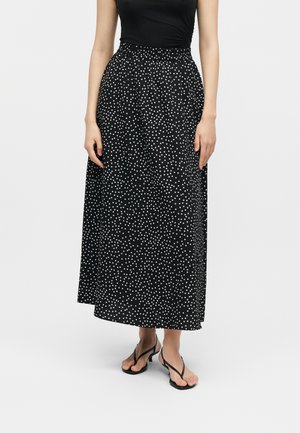 Žena v dlhom čierno-bielom bodkovanom sukni, čiernom tielku bez rukávov a čiernych sandáloch so zvrškami stojí na jednolrom pozadí.