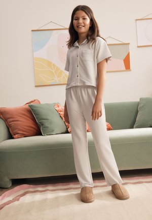 Pige står i lysegrå stribede pyjamas og hjemmesko i en stue med en grøn sofa og abstrakt vægkunst.