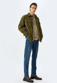 Chaqueta de botonera verde oliva con dos bolsillos en el pecho, camiseta crema debajo, jeans azules y botas marrones hasta el tobillo. Texturas suaves en toda la prenda.