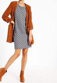 Giacca in suede marrone sopra un abito a fantasia bianco e nero, caratterizzato da un design geometrico. Stivaletti alla caviglia color nocciola completano l'outfit.