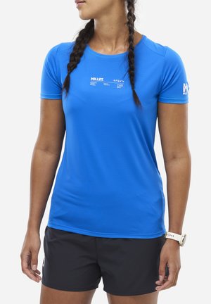 Femme aux cheveux tressés portant une chemise à manches courtes bleue Millet, un short noir et une montre blanche au poignet, vue de profil.