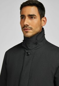 Zwarte jas met een hoge kraag, voorzien van twee knoopsluitingen en een textuurstof. Geen patronen of versieringen.