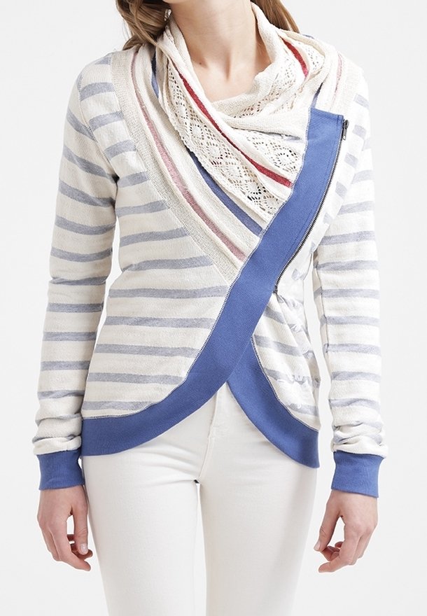 Femme portant un pull zippé asymétrique rayé avec une bordure bleue, superposé à un haut orné de dentelle et un pantalon blanc.