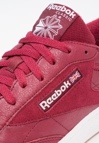 Bordové klasické tenisky Reebok, které kombinují kůži a semiš, mají texturované šňůrky a logo s detailem britské vlajky.