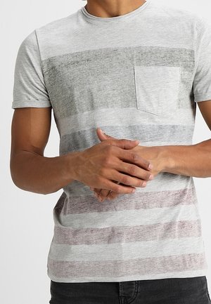 Man draagt een lichtgrijs gestreept T-shirt met korte mouwen en een klein borstzakje, handen samengevouwen voor de taille.