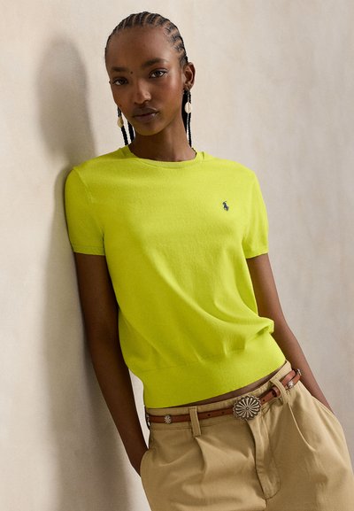 Πλεκτό πουλόβερ με κοντά μανίκια σε φωτεινό νέον κίτρινο, με ριμπ λαιμόκοψη και μικρό λογότυπο στο στήθος. Συνδυασμένο με μπεζ παντελόνια.