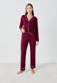 Bordeaux pyjama set met lange mouwen en een broek, voorzien van witte bies langs de kraag en manchetten. Gladde stof en relaxte pasvorm.